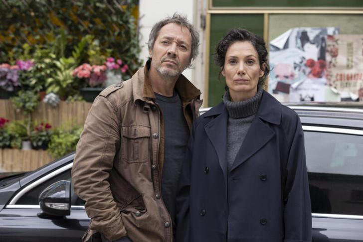 Bruno Debrandt et Maëlle Mietton, adossés à la voiture de la commissaire, regardant fixement la devanture d'une librairie (qu'on ne voit pas)